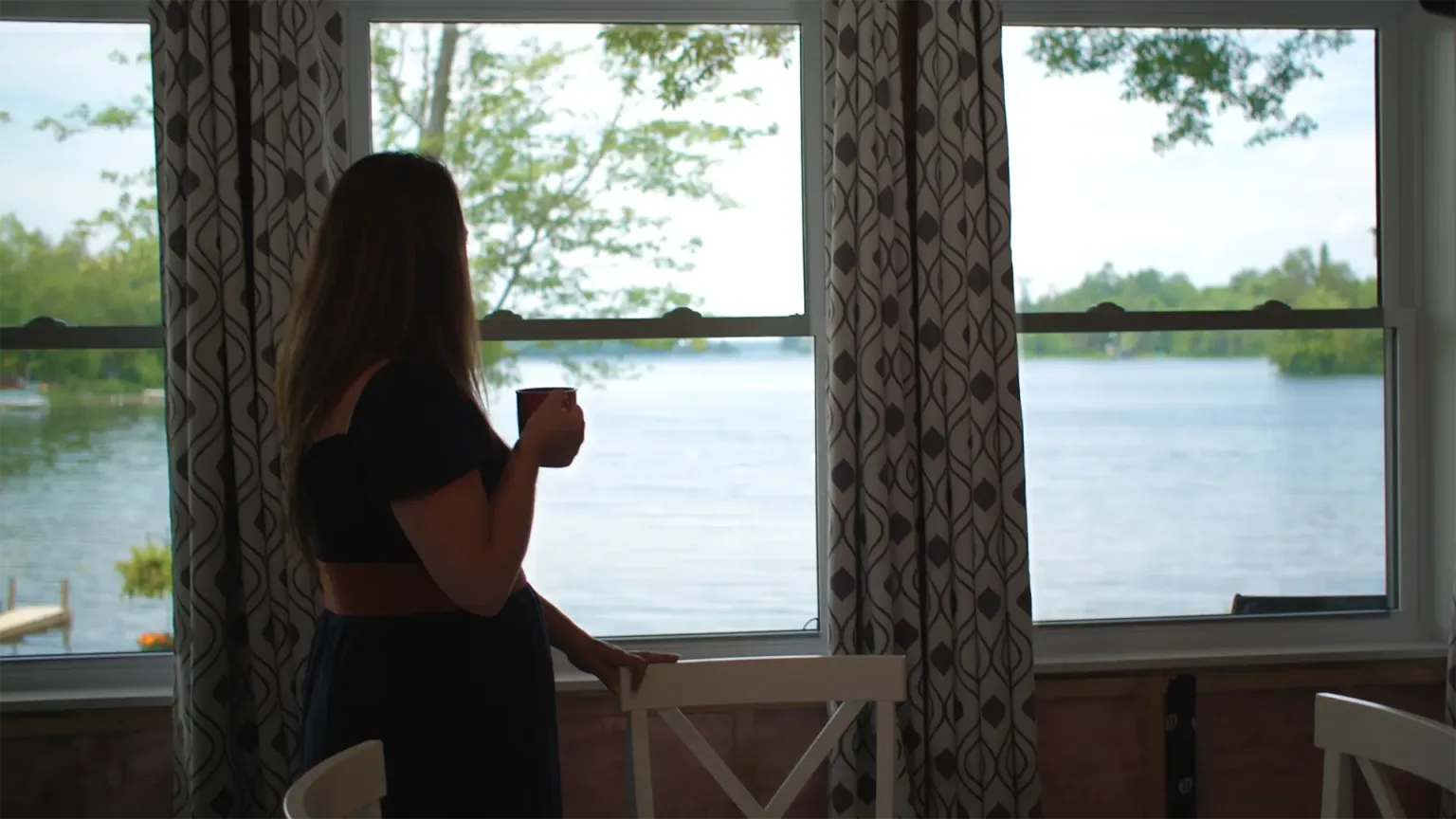 A woman stands by a window, looking at a lake with lush greenery in the background