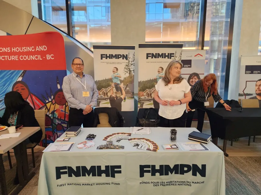 A man and a woman Senior Program Officers (SPOs) standing behind an informational booth table for the First Nations Market Housing Fund (FNMHF) meeting with guests at an indoor event.